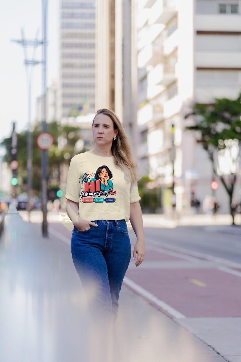 May include: A cream-colored short-sleeve t-shirt featuring a graphic print of a person with dark hair, the text "HI Ask me anything", and three small icons labeled "TEXT", "VOICE", and "IMAGE" in red, green, and blue rectangular boxes.
