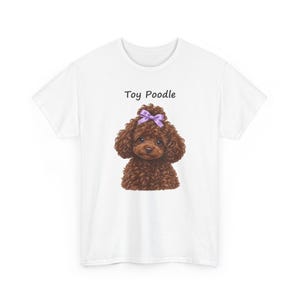 May include: A white short-sleeved t-shirt featuring a graphic of a brown curly-haired toy poodle with a purple bow on its head. The text Toy Poodle is printed in black above the dog illustration, which is centered on the front of the shirt.