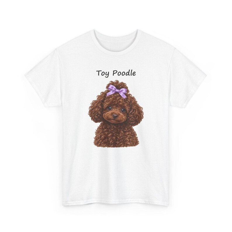 May include: A white short-sleeved t-shirt featuring a graphic of a brown curly-haired toy poodle with a purple bow on its head. The text Toy Poodle is printed in black above the dog illustration, which is centered on the front of the shirt.