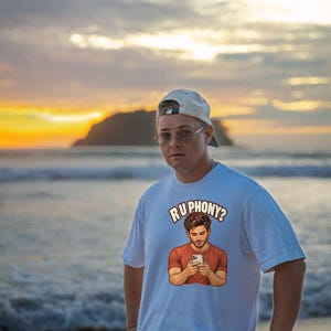 May include: A white short-sleeved t-shirt featuring a graphic print of a person looking at a smartphone with the text "RU PHONY?" arched above the illustration. The shirt has a crew neckline and a relaxed fit, made of a solid white fabric.