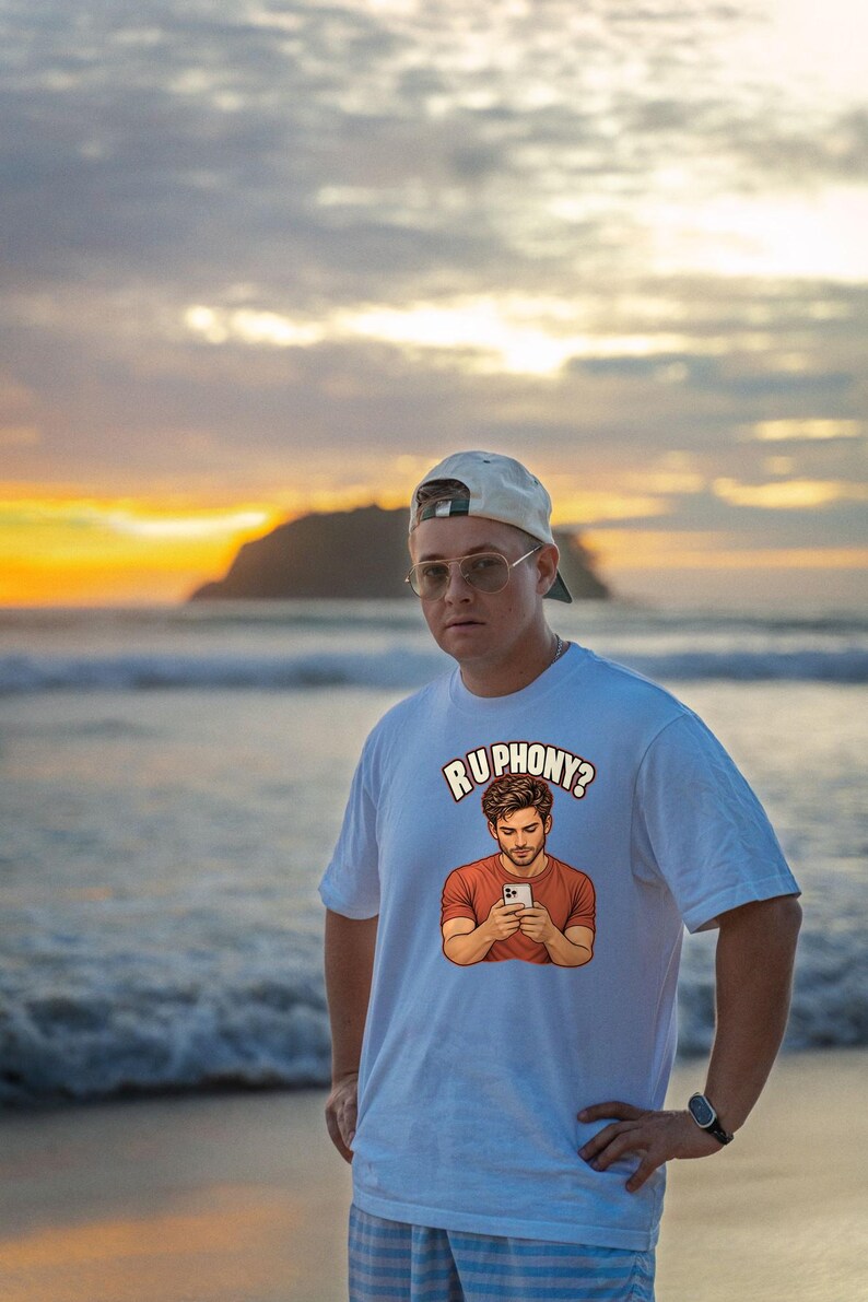 May include: A white short-sleeved t-shirt featuring a graphic print of a person looking at a smartphone with the text "RU PHONY?" arched above the illustration. The shirt has a crew neckline and a relaxed fit, made of a solid white fabric.