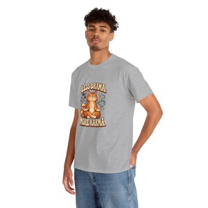 May include: A heather gray short-sleeve t-shirt featuring a graphic of an orange tabby cat sitting in a lotus position. The text above the cat reads LESS DRAMA and the text below reads MORE KARMA in a bold, retro-style font.