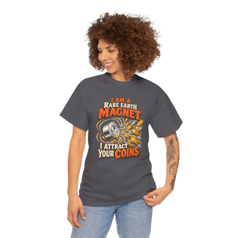 May include: A charcoal gray t-shirt featuring a graphic of a smiling cylindrical magnet attracting various gold and silver coins. The text reads, I AM A RARE EARTH MAGNET I ATTRACT YOUR COINS, printed in bold orange and white lettering on the front.