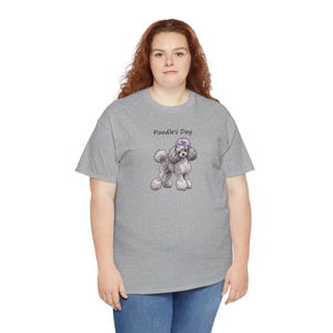 May include: A heather gray short-sleeve t-shirt featuring a graphic of a gray poodle with a purple bow on its head. Above the poodle, the text Poodle's Day is printed in a simple black font. The shirt has a crew neckline and a standard fit.