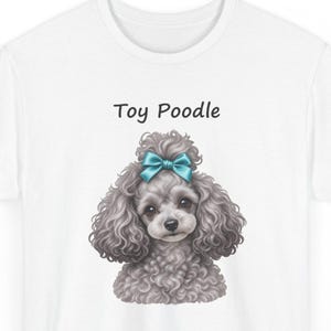K&ouml;nnte beinhalten: Ein wei&szlig;es T-Shirt mit einer grafischen Illustration eines grauen Toypudels mit lockigem Fell und einer t&uuml;rkisfarbenen Schleife auf dem Kopf. Der Text "Toy Poodle" ist in schwarzen Buchstaben &uuml;ber dem Kopf des Hundes aufgedruckt.