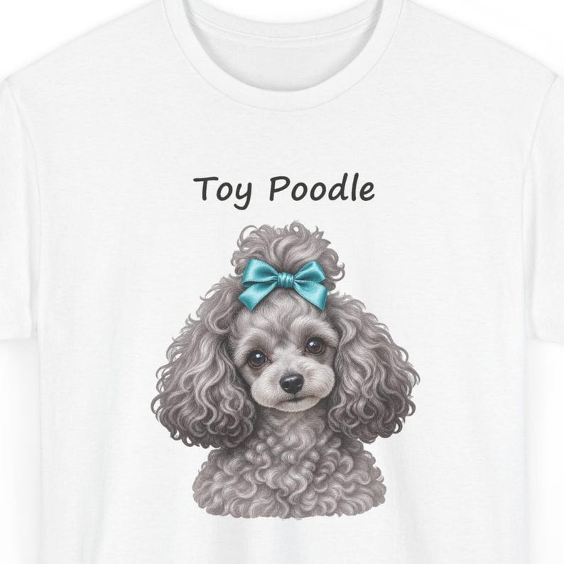 K&ouml;nnte beinhalten: Ein wei&szlig;es T-Shirt mit einer grafischen Illustration eines grauen Toypudels mit lockigem Fell und einer t&uuml;rkisfarbenen Schleife auf dem Kopf. Der Text "Toy Poodle" ist in schwarzen Buchstaben &uuml;ber dem Kopf des Hundes aufgedruckt.