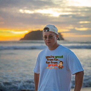 May include: A white short-sleeved t-shirt featuring a graphic print with the text you're great from far... far from great. The design includes a cartoon illustration of a person using binoculars and a row of small orange footprints.
