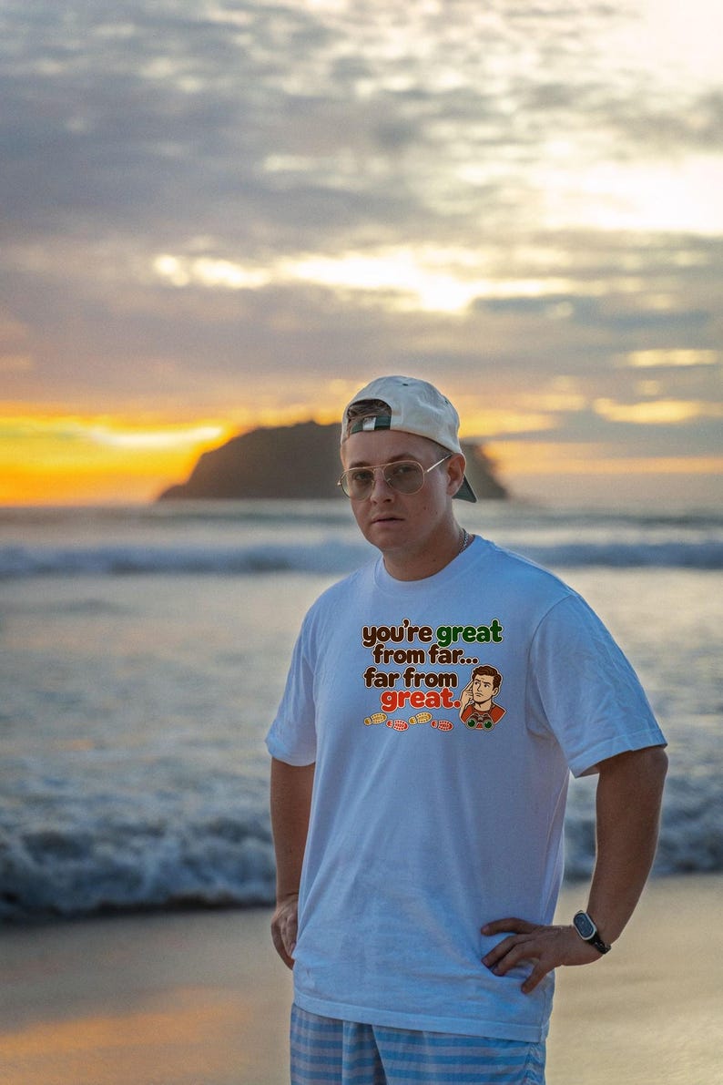 May include: A white short-sleeved t-shirt featuring a graphic print with the text you're great from far... far from great. The design includes a cartoon illustration of a person using binoculars and a row of small orange footprints.