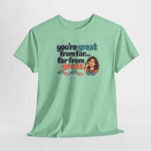 May include: A light green short-sleeve t-shirt featuring a graphic of a person with binoculars and the text: you're great from far... far from great. Below the text are three small colorful footprints in blue, red, and orange.