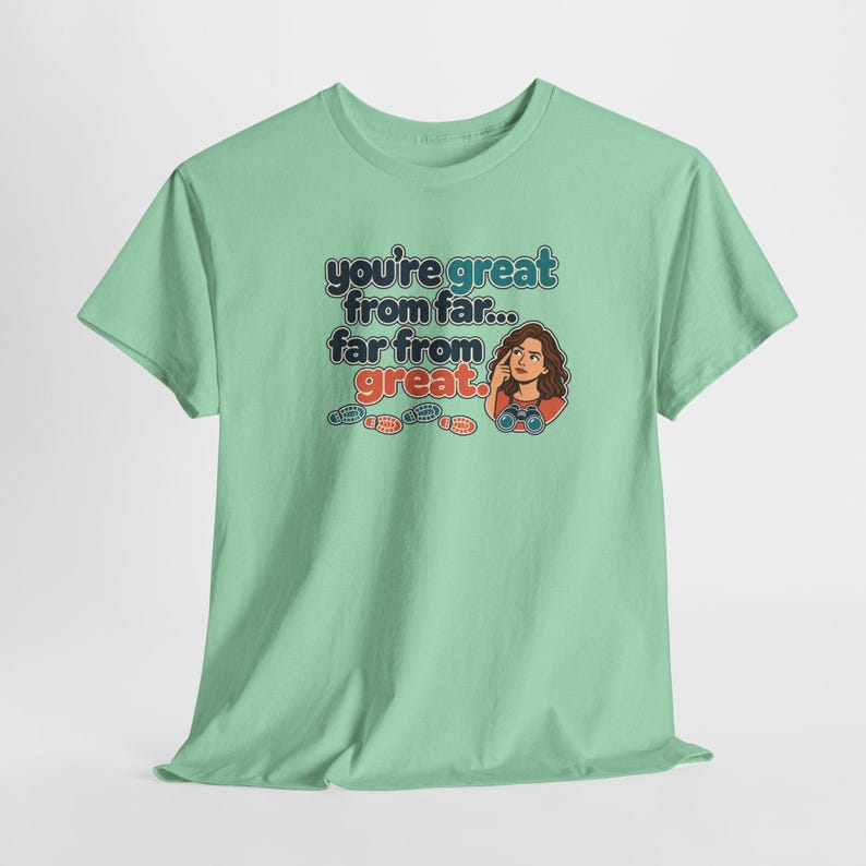 May include: A light green short-sleeve t-shirt featuring a graphic of a person with binoculars and the text: you're great from far... far from great. Below the text are three small colorful footprints in blue, red, and orange.