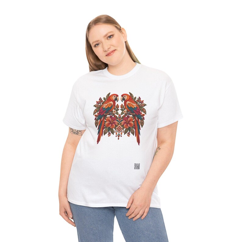 May include: A white short-sleeve t-shirt featuring a central graphic of two symmetrical parrots perched among orange hibiscus flowers and green leaves. The design includes intricate patterns and a small QR code near the bottom hem.
