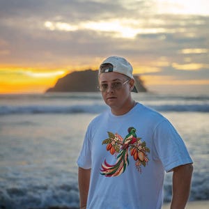May include: A white short-sleeved t-shirt featuring a colorful graphic print of a quetzal bird perched on a branch with cacao pods and white flowers. The bird has vibrant green, red, and yellow plumage with a long, flowing tail.