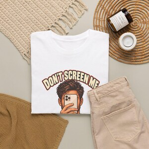 Op de afbeelding: Een wit t-shirt met een afbeelding van een persoon die een smartphone voor het gezicht houdt, met daarboven de tekst "DON'T SCREEN ME" in een boog, geschreven in een vet retro-lettertype met een donkere rand.