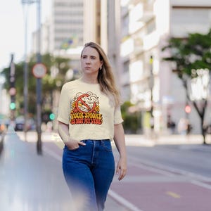 May include: A cream-coloured short-sleeve t-shirt featuring a graphic print of a cigarette inside a red prohibition circle with smoke clouds and orange flowers. The text reads YOUR SMOKE IS NOT MY VIBE in a bold, retro-style orange and red font.