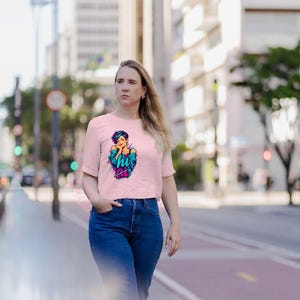 Pu&ograve; includere: Una t-shirt a maniche corte color rosa chiaro con una stampa grafica di una persona con capelli corti blu che indossa una giacca color ottanio. La grafica include il testo "hi" in un carattere stilizzato. Abbinata a jeans blu scuro.