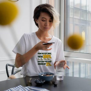 May include: A white short-sleeved t-shirt featuring the black and yellow text: FROM 100 FEET AWAY, YOU'RE 100%. SADLY, EVERY FOOT CLOSER KNOCKS OFF 1%. The graphic includes a small horizontal line with tick marks and arrows.