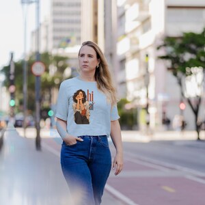 May include: A light blue short-sleeved t-shirt featuring a graphic illustration of a person with dark hair wearing a rust-colored top and an unbuttoned shirt, accompanied by the text "hi ai" printed in a vertical orange font.