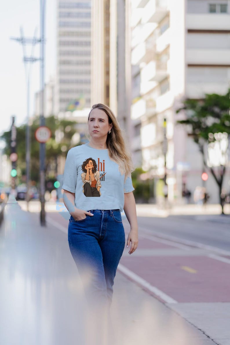 May include: A light blue short-sleeved t-shirt featuring a graphic illustration of a person with dark hair wearing a rust-colored top and an unbuttoned shirt, accompanied by the text "hi ai" printed in a vertical orange font.