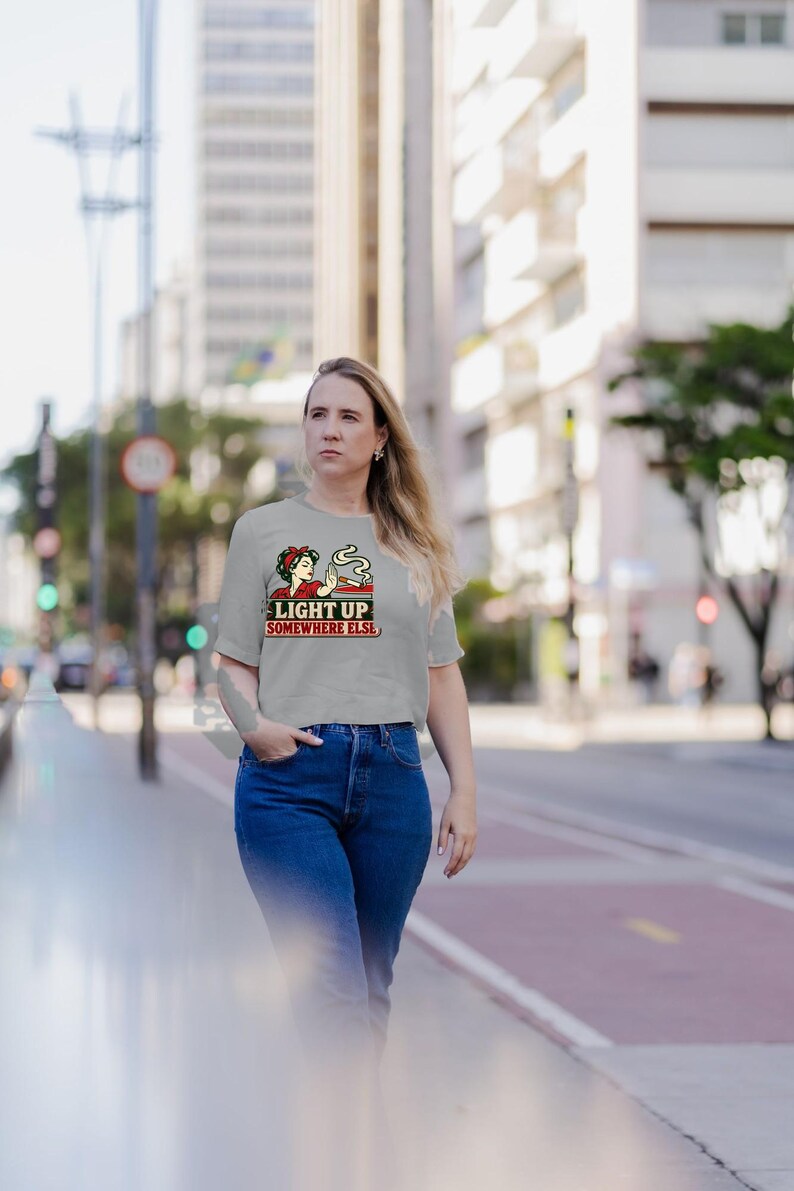 May include: A light gray short-sleeved crop top featuring a graphic illustration of a person with a red headband holding a lit cigarette. The text "LIGHT UP SOMEWHERE ELSE" is printed in bold red letters below the illustration on the front.