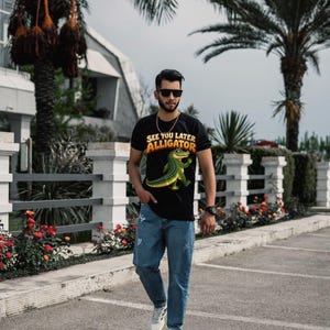 May include: A black short-sleeved t-shirt featuring a graphic of a green alligator and the text "SEE YOU LATER ALLIGATOR" in yellow and orange, paired with blue denim jeans and white athletic sneakers with black stripes.