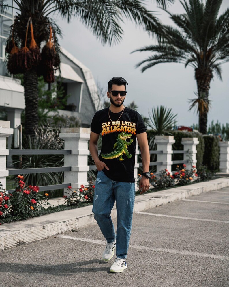 May include: A black short-sleeved t-shirt featuring a graphic of a green alligator and the text "SEE YOU LATER ALLIGATOR" in yellow and orange, paired with blue denim jeans and white athletic sneakers with black stripes.