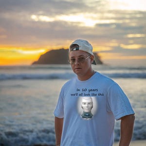 May include: A white short-sleeve t-shirt featuring a graphic of a robotic head and the text "In 10 years we'll all look like this" printed on the chest. The shirt is worn with a light-colored baseball cap and wire-rimmed glasses.