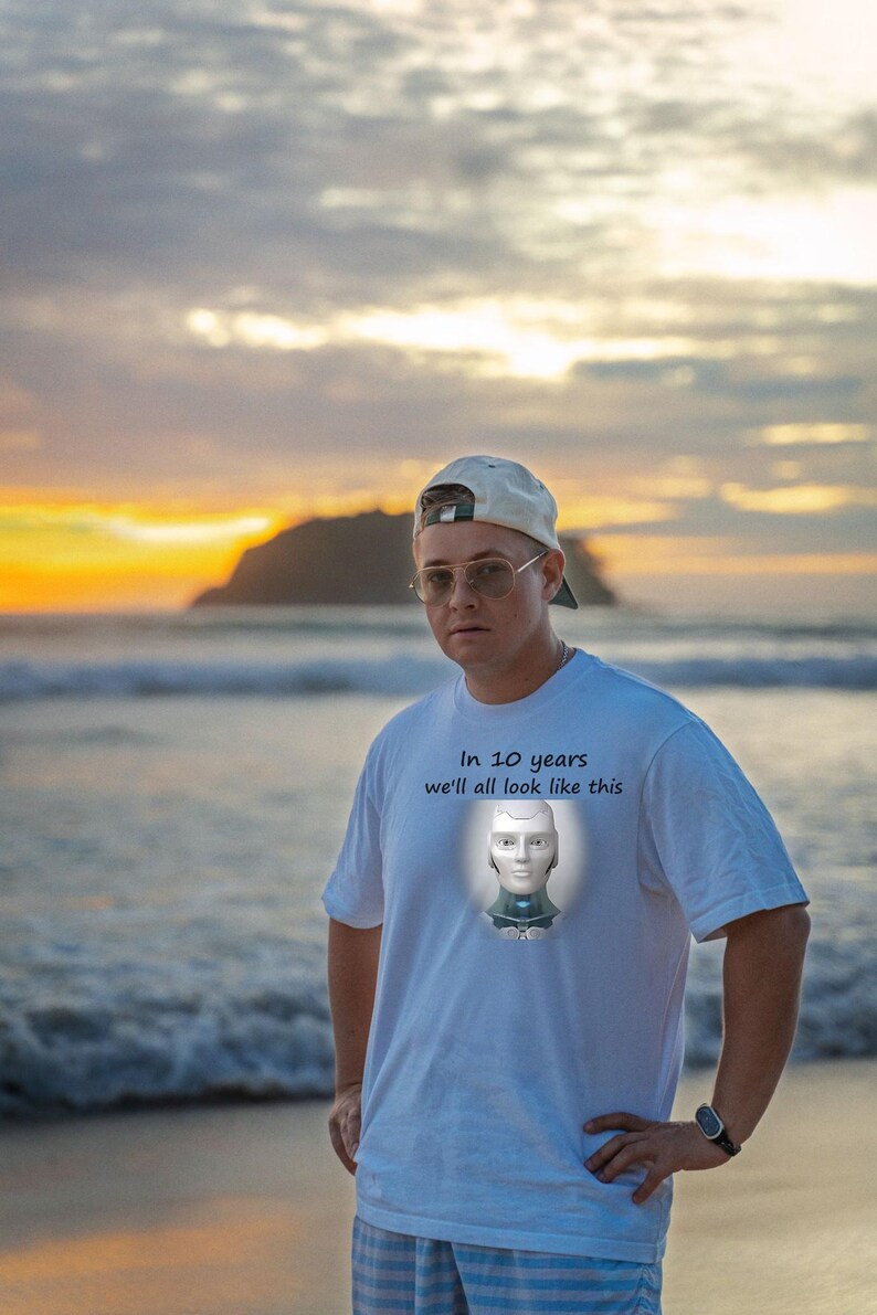 May include: A white short-sleeve t-shirt featuring a graphic of a robotic head and the text "In 10 years we'll all look like this" printed on the chest. The shirt is worn with a light-colored baseball cap and wire-rimmed glasses.