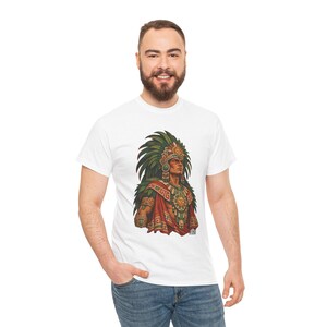 May include: A white short-sleeved t-shirt featuring a graphic print of an Aztec warrior wearing a large green feathered headdress, a gold sun medallion, and red and green patterned clothing. The illustration is centered on the chest.