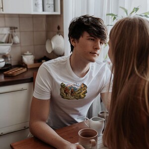 Puede incluir: Una camiseta blanca de manga corta con un estampado gr&aacute;fico rectangular en el pecho. El gr&aacute;fico muestra una escena de paisaje detallada con dos figuras caminando por un sendero hacia monta&ntilde;as nevadas bajo un cielo azul.