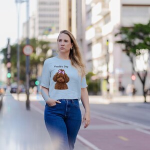 May include: A light blue short-sleeved t-shirt featuring a graphic of a brown poodle with a pink bow on its head and the text Poodle's Day printed above the dog. The shirt is paired with dark blue denim jeans.