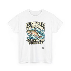 May include: A white t-shirt featuring a graphic of a fish jumping through blue waves. The text above the fish reads CURRENTLY SWIMMING and the text below reads AGAINST THE CURRENT. A small square QR code is printed near the bottom right.