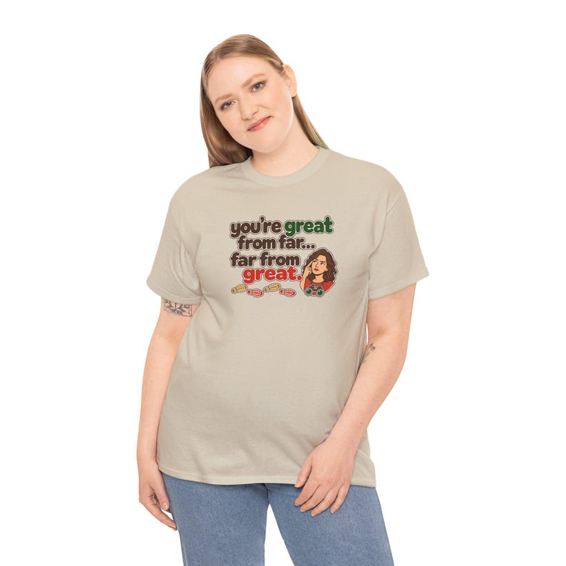 May include: A beige short-sleeve t-shirt featuring the text "you're great from far... far from great." in brown, green, and red lettering, accompanied by a small illustration of a person holding a game controller.