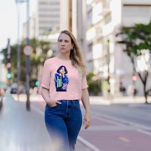 Puede incluir: Una camiseta de manga corta de color rosa claro con una ilustraci&oacute;n gr&aacute;fica de una persona de pelo oscuro con un su&eacute;ter azul, con el texto "hi" en verde claro y "ai" en blanco impreso bajo la ilustraci&oacute;n, combinada con vaqueros azules.