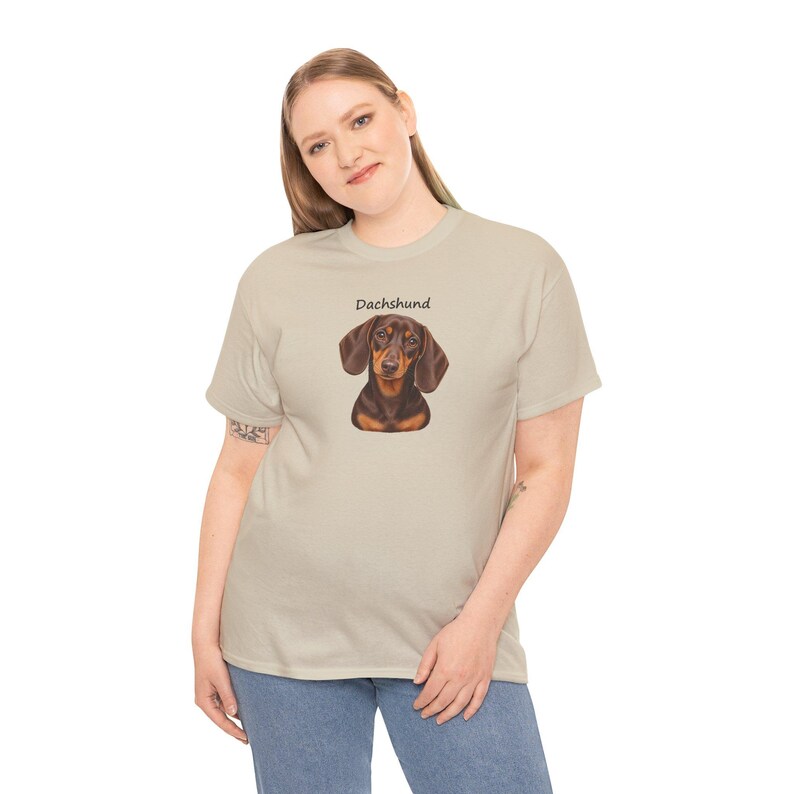 K&ouml;nnte beinhalten: Ein beiges Kurzarm-T-Shirt mit einer Grafik eines braun-schwarzen Dackels, die mittig auf der Brust platziert ist. &Uuml;ber der Hundeillustration ist das Wort Dachshund in einer einfachen schwarzen serifenlosen Schrift gedruckt.