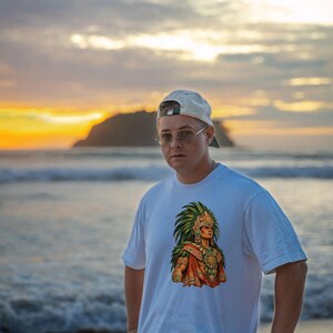 May include: A white short-sleeved t-shirt featuring a colorful graphic print of an individual wearing an elaborate green feathered headdress and traditional ornate jewelry, centered on the chest of the garment.