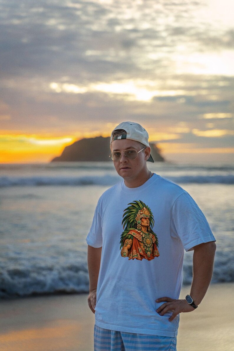 May include: A white short-sleeved t-shirt featuring a colorful graphic print of an individual wearing an elaborate green feathered headdress and traditional ornate jewelry, centered on the chest of the garment.