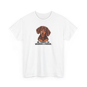 May include: A white short-sleeved t-shirt featuring a brown and black dachshund illustration centered on the chest. Below the dog, the text reads: HE THINKS I'M WEARING A MIRROR. The graphic is printed in black, bold, sans-serif capital letters.