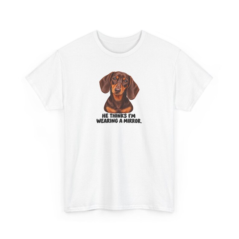 May include: A white short-sleeved t-shirt featuring a brown and black dachshund illustration centered on the chest. Below the dog, the text reads: HE THINKS I'M WEARING A MIRROR. The graphic is printed in black, bold, sans-serif capital letters.