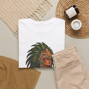 May include: A white t-shirt featuring a graphic print of a person wearing a large, ornate green and red feathered headdress with a central sun emblem. The person has detailed facial features and decorative jewelry around the neck and ears.