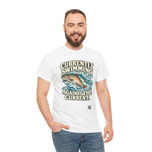May include: A white short-sleeved t-shirt featuring a graphic of a fish jumping through blue waves. The text reads "CURRENTLY SWIMMING AGAINST THE CURRENT." The design includes a small black square icon near the bottom of the graphic.