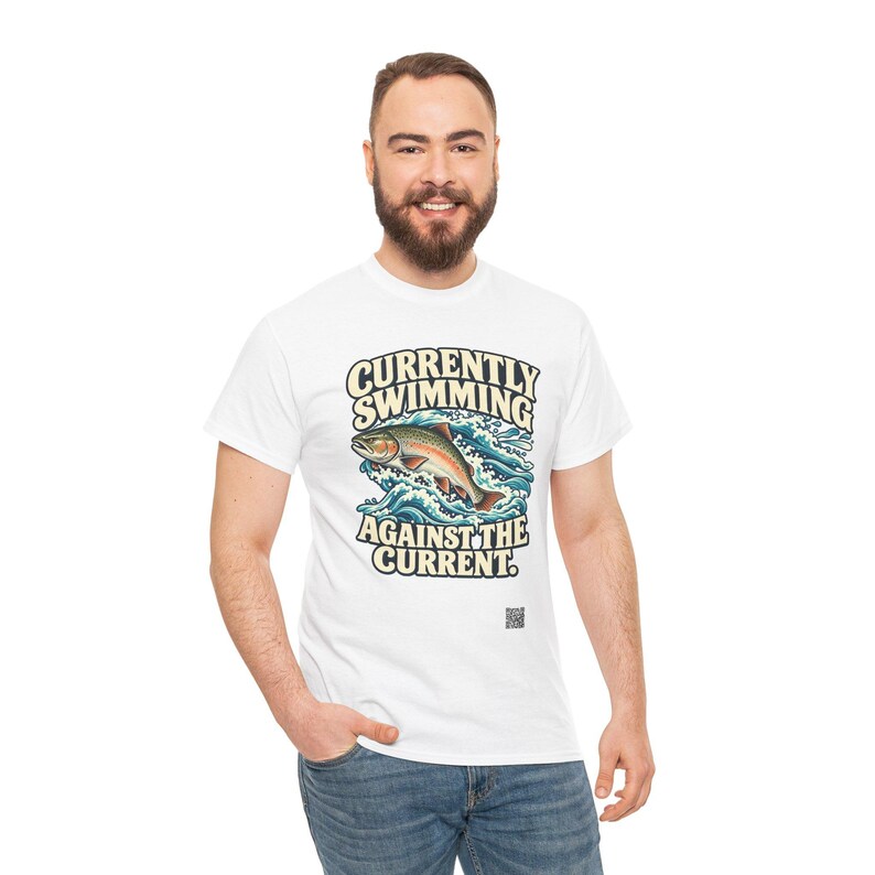 May include: A white short-sleeved t-shirt featuring a graphic of a fish jumping through blue waves. The text reads "CURRENTLY SWIMMING AGAINST THE CURRENT." The design includes a small black square icon near the bottom of the graphic.