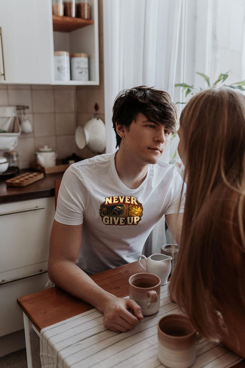 Puede incluir: Una camiseta blanca de manga corta con un gr&aacute;fico central que muestra el texto NEVER GIVE UP en letras doradas, acompa&ntilde;ado de la ilustraci&oacute;n de una peque&ntilde;a figura extrayendo oro dentro de un marco rocoso y texturizado.