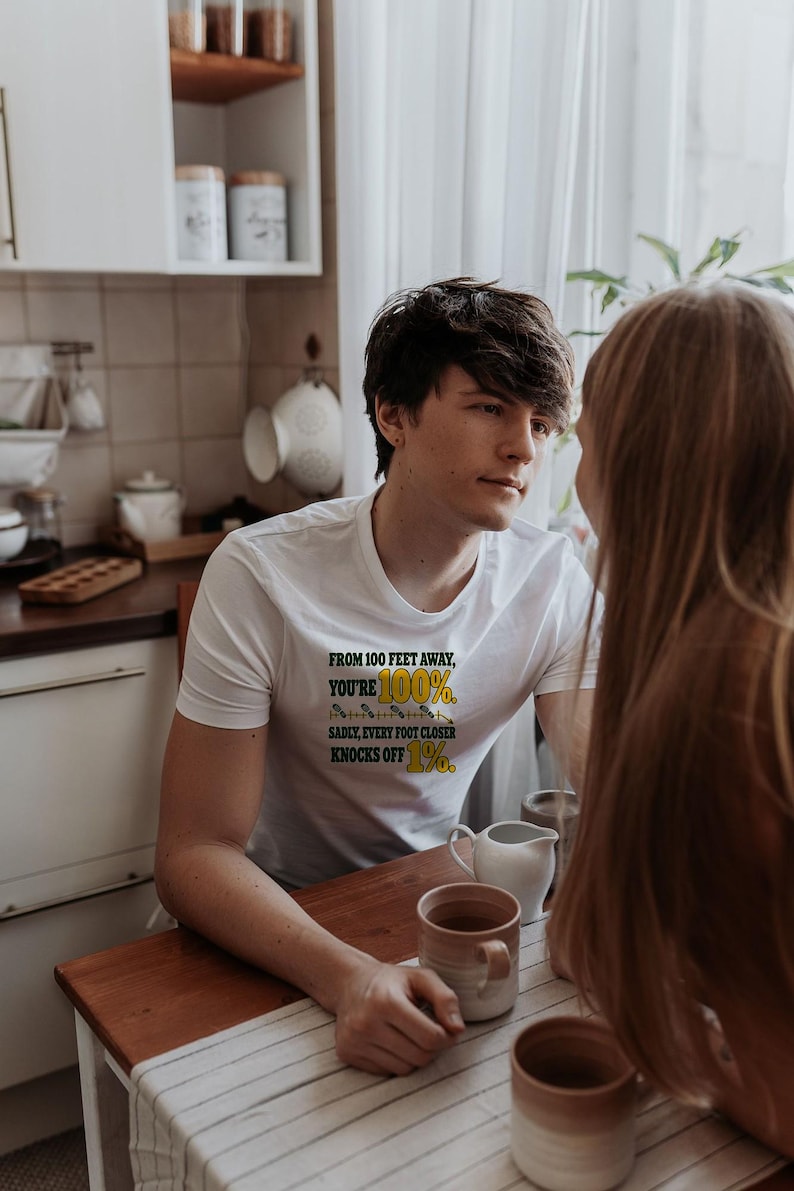 May include: A white short-sleeved t-shirt featuring the text "FROM 100 FEET AWAY, YOU'RE 100%. SADLY, EVERY FOOT CLOSER KNOCKS OFF 1%." The text is printed in black and yellow, with a small graphic of a measuring tape between the lines.