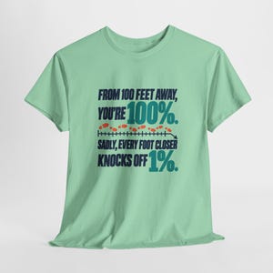 May include: A light green short-sleeve t-shirt featuring dark blue and teal text that reads: FROM 100 FEET AWAY, YOU'RE 100%. SADLY, EVERY FOOT CLOSER KNOCKS OFF 1%. The design includes a horizontal line with small orange footprints above it.
