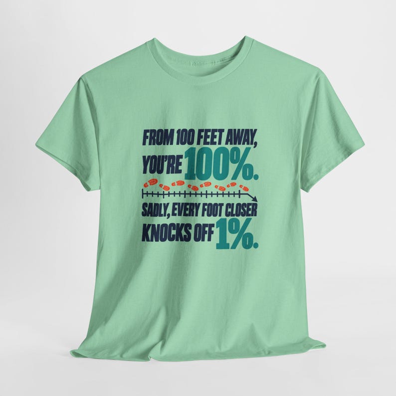 May include: A light green short-sleeve t-shirt featuring dark blue and teal text that reads: FROM 100 FEET AWAY, YOU'RE 100%. SADLY, EVERY FOOT CLOSER KNOCKS OFF 1%. The design includes a horizontal line with small orange footprints above it.