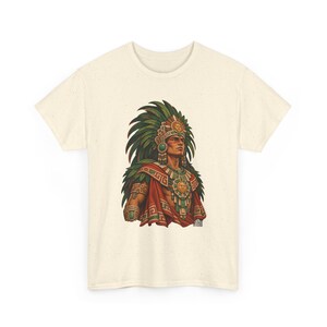 May include: A cream-colored short-sleeve t-shirt featuring a graphic print of a person wearing a large green feathered headdress, a red cape, and ornate gold jewelry with sun motifs. The design includes intricate geometric patterns.