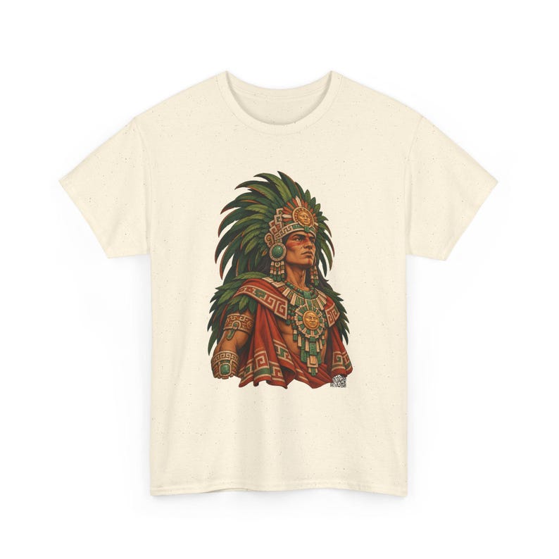 May include: A cream-colored short-sleeve t-shirt featuring a graphic print of a person wearing a large green feathered headdress, a red cape, and ornate gold jewelry with sun motifs. The design includes intricate geometric patterns.