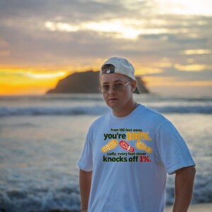May include: A white short-sleeved t-shirt featuring the text 'from 100 feet away, you're 100%. sadly, every foot closer knocks off 1%.' along with three colorful shoe print graphics in yellow, red, and yellow.