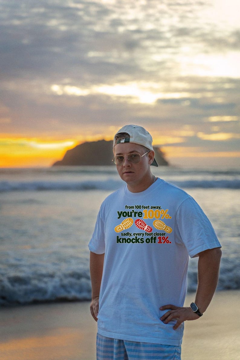 May include: A white short-sleeved t-shirt featuring the text 'from 100 feet away, you're 100%. sadly, every foot closer knocks off 1%.' along with three colorful shoe print graphics in yellow, red, and yellow.