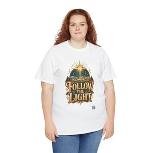 May include: A white short-sleeved t-shirt featuring a graphic print of a stylized landscape with a bright star above a town. The text "FOLLOW THE LIGHT" is printed in large, gold-toned letters below the illustration, with a small QR code at the bottom.